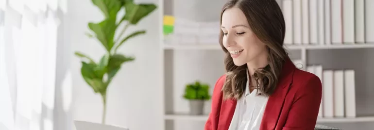 Smiling woman in a red blazer working on a laptop in a bright office.