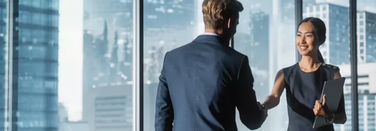 Two business professionals shaking hands in a glass-walled office with a cityscape view.