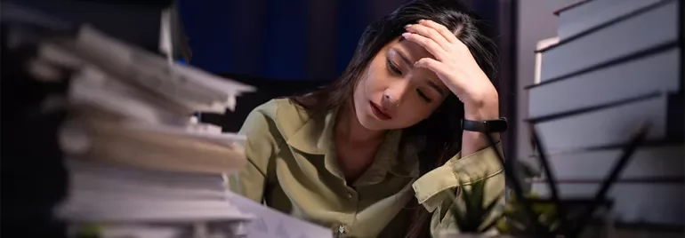 A stressed lady sitting with her hand on her head in a dark office with piles of books and papers on either side of her desk