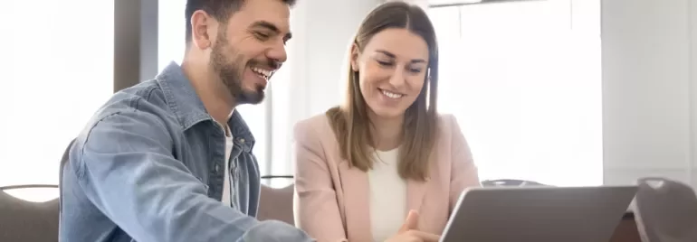 Two people are sitting at a table facing a laptop while smiling.