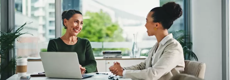 Two women engaged in discussion at a table with a laptop open between them