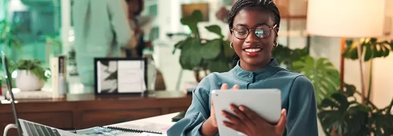 Woman standing in the office on her ipad with her laptop behind her