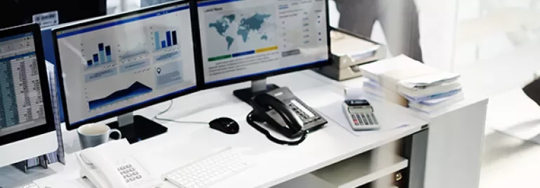 Office desk with dual monitors displaying charts and data, phone, and keyboard.