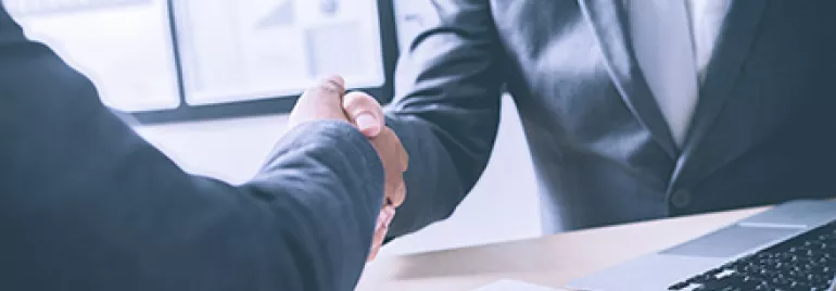 Two businesspeople shaking hands at a desk with documents and a laptop.