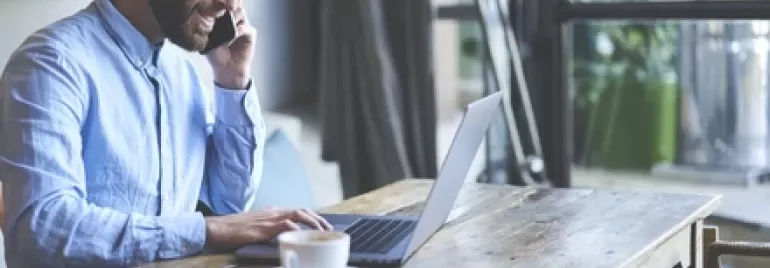 Man talking on phone while working on laptop at a wooden table.