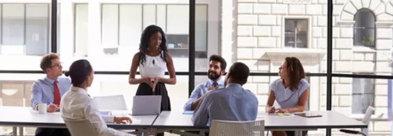 A woman stands and speaks to a group in an office meeting room.