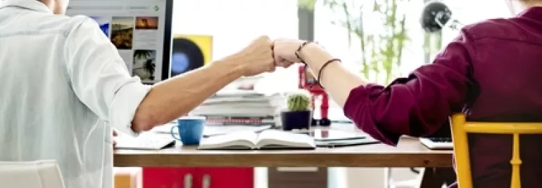Two people fist bumping at a shared desk with a computer.