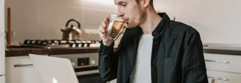 Man drinking coffee while working on a laptop in a kitchen.