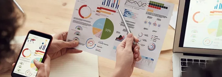 Person analyzing charts on paper with phone and laptop at desk.