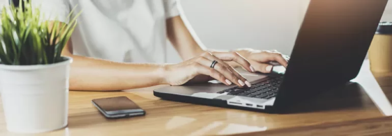 Person typing on a laptop at a wooden desk with a plant and smartphone nearby.