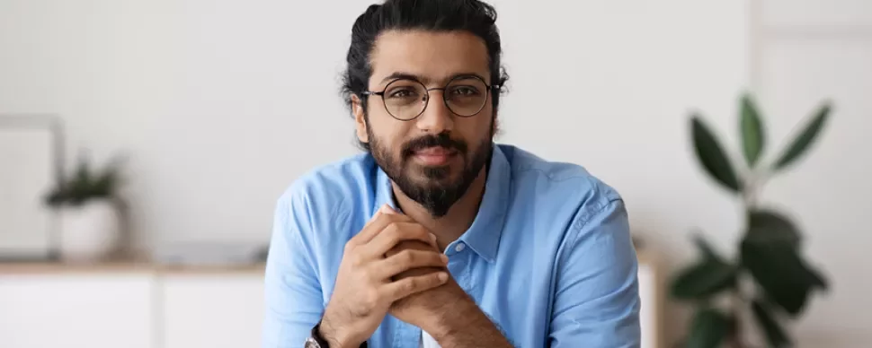 Man with glasses and beard in a blue shirt, sitting indoors.