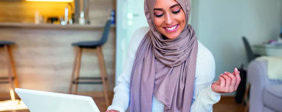 Woman in a hijab smiling at a laptop in a modern living space.