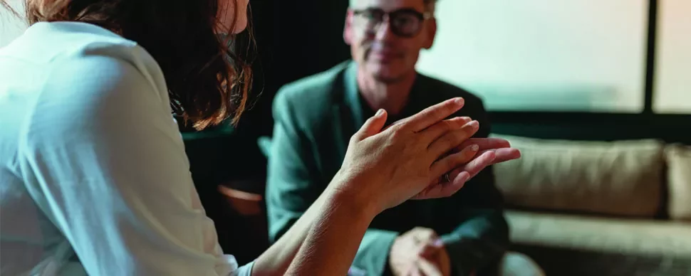 Close-up of hands gesturing during a conversation, with a person sitting on a sofa in the background.