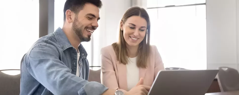 Two people are sitting at a table facing a laptop while smiling.