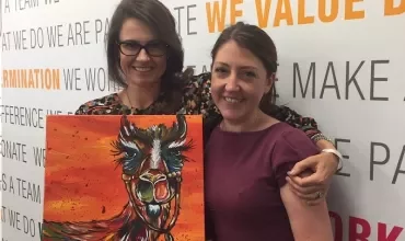 Two women smiling, holding a colorful abstract painting of an animal.