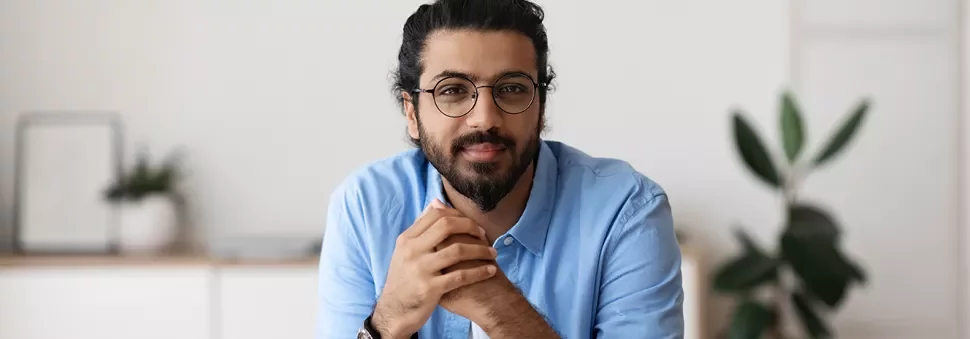 Man with glasses and beard in a blue shirt, sitting indoors.