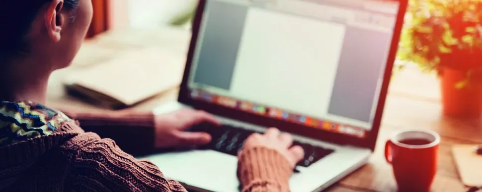 A person typing on a laptop with a blank document on the screen, a notepad, and a coffee cup on the wooden table.