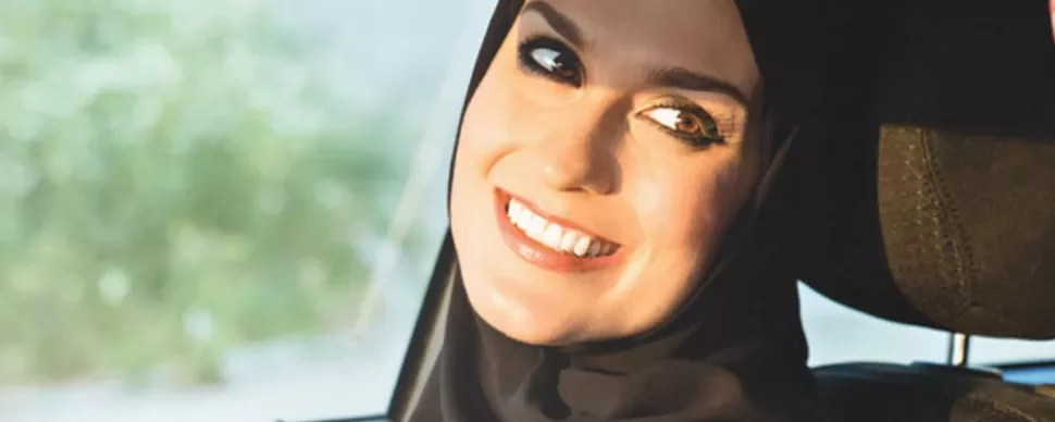 Smiling person wearing a hijab in a car seat, sunlight on face.
