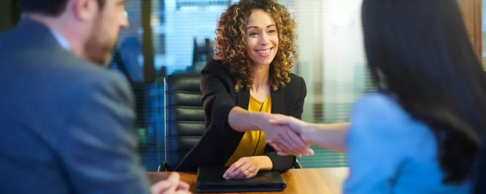 Woman in business attire smiling and shaking hands across a desk.