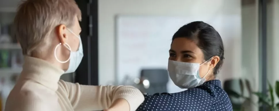 Two women wearing masks greet each other with an elbow bump.