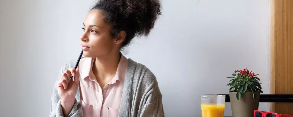Woman in a sweater holding a pen and looking thoughtful, with juice and plant nearby.
