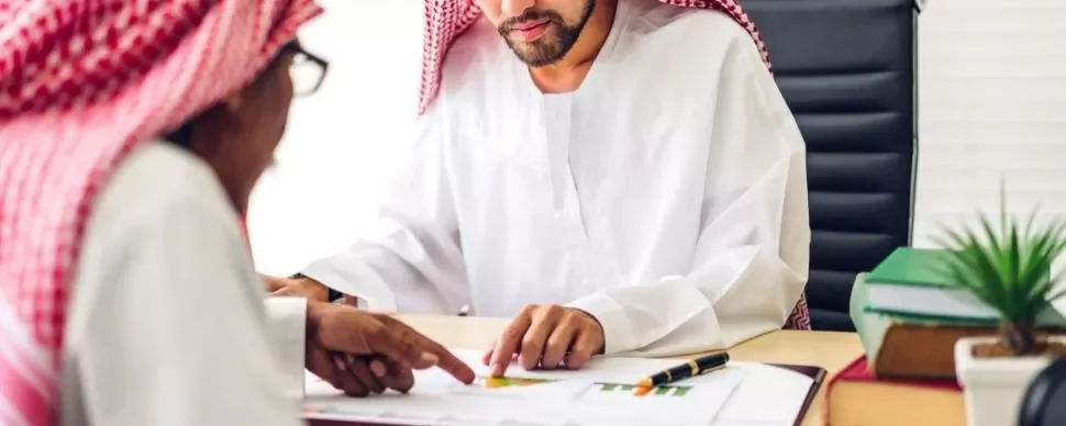 Two men in traditional attire reviewing documents at a desk.