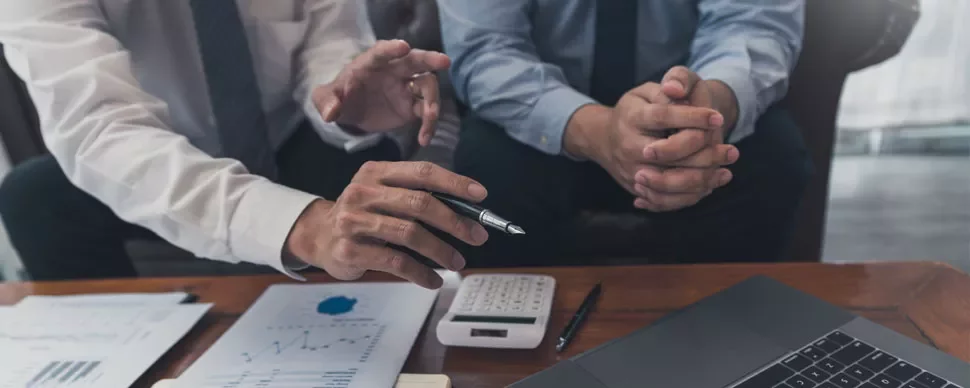 Two people discussing charts with a pen and calculator on a table.