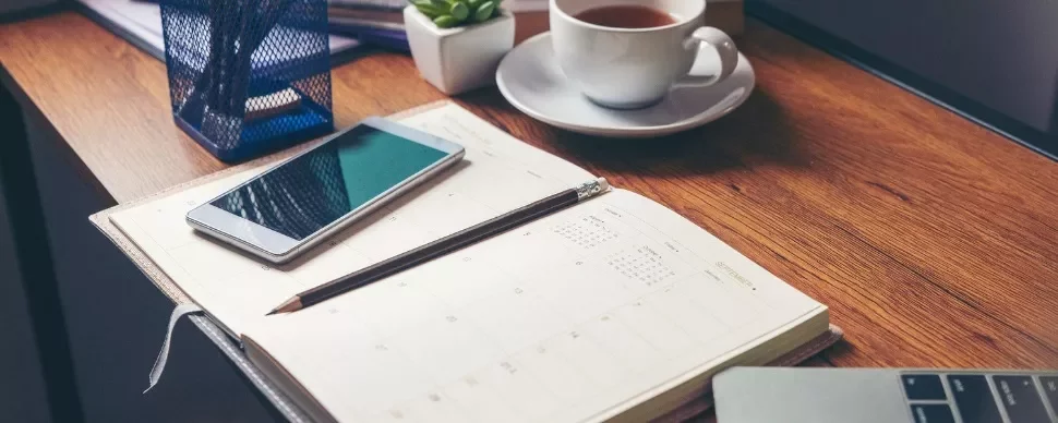 A wooden desk with a planner, smartphone, pencil, coffee cup, and a small plant.