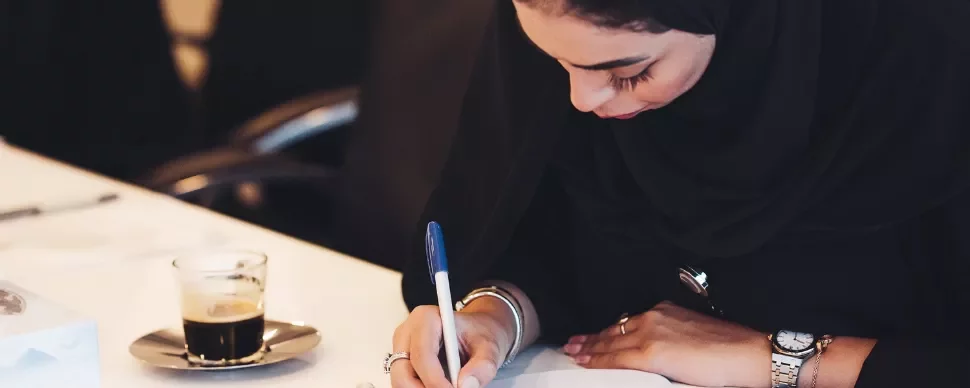 Woman in hijab writing at a desk with a cup of coffee nearby.
