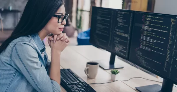 Woman in glasses focused on coding at dual monitors.
