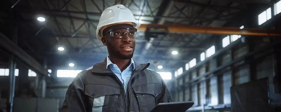 Engineer in a hard hat and safety glasses holding a tablet in a factory.