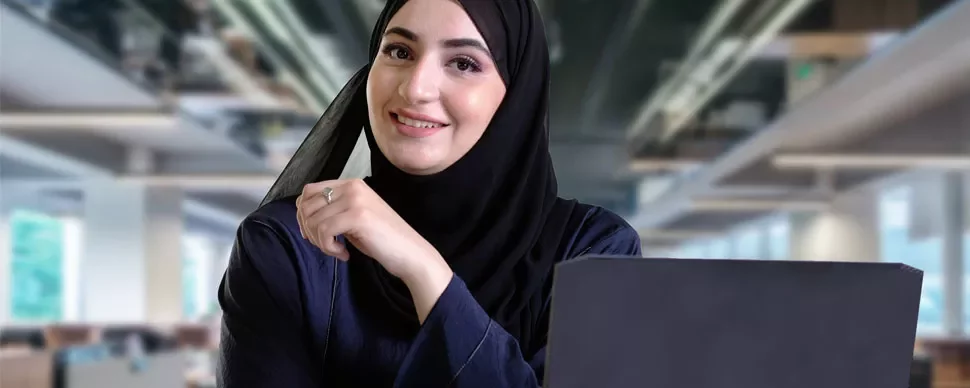 Smiling woman in hijab working on a laptop in an office.