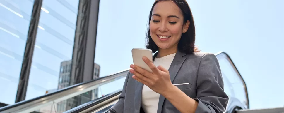Woman in a suit smiling at her phone while going up an escalator outdoors.