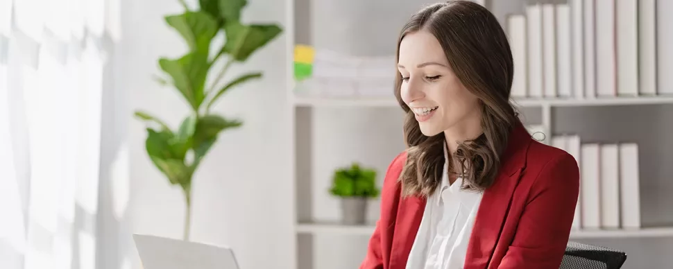 Smiling woman in a red blazer working on a laptop in a bright office.