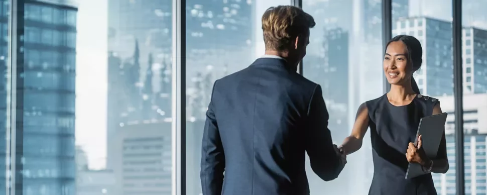 Two business professionals shaking hands in a glass-walled office with a cityscape view.
