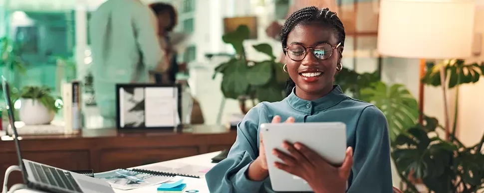 Woman standing in the office on her ipad with her laptop behind her