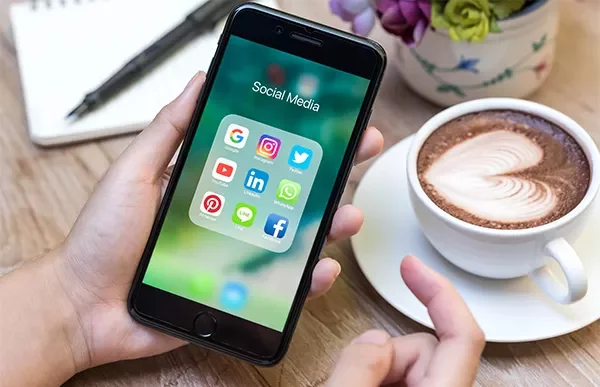 Person holding a smartphone displaying social media apps next to a latte with heart art.
