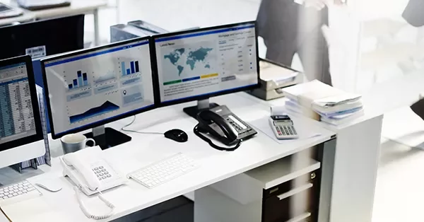 Office desk with dual monitors displaying charts and data, phone, and keyboard.