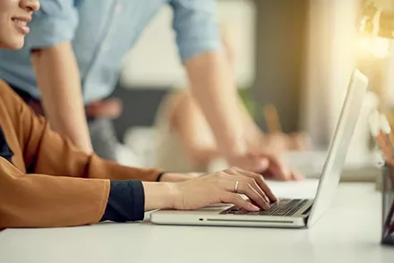 Person typing on a laptop at a desk with another person standing nearby.