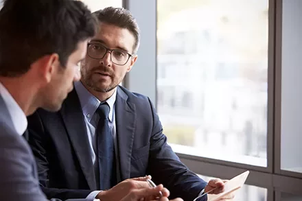 Two businessmen in a meeting, one holding a document, sitting near a window.