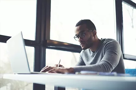 Man wearing glasses working on a laptop near large windows.