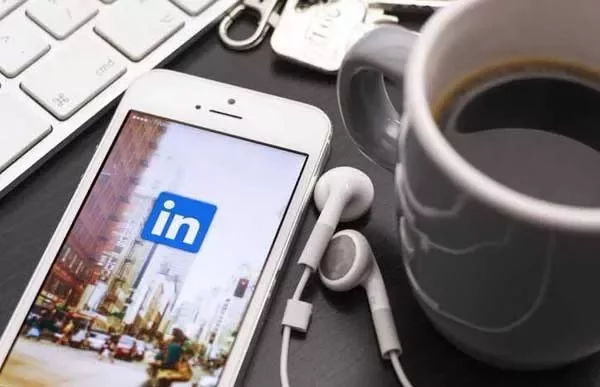 Smartphone with LinkedIn app on screen, beside earbuds, keyboard, and coffee mug.