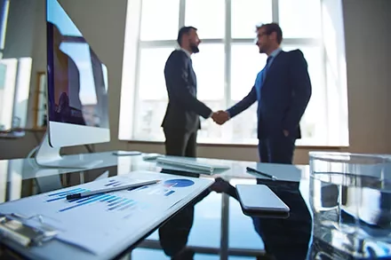 Two businessmen shaking hands in an office with a desktop and charts visible.