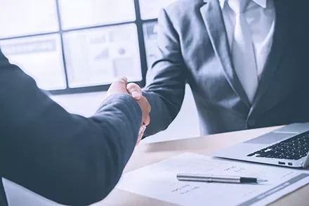 Two businesspeople shaking hands at a desk with documents and a laptop.