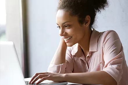 Smiling woman working on a laptop near a window.