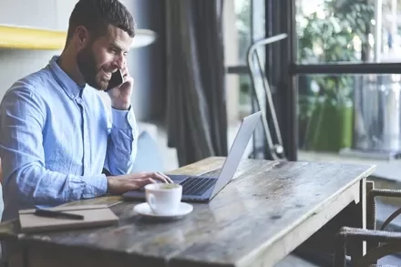 Man talking on phone while working on laptop at a wooden table.