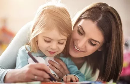 Smiling woman helps young girl draw with a pen.