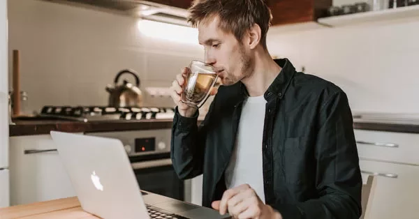 Man drinking coffee while working on a laptop in a kitchen.