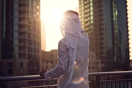 A person in traditional attire stands on a balcony overlooking a cityscape at sunrise.