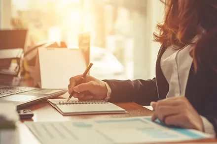 Person writing notes at a desk with sunlight in the background.